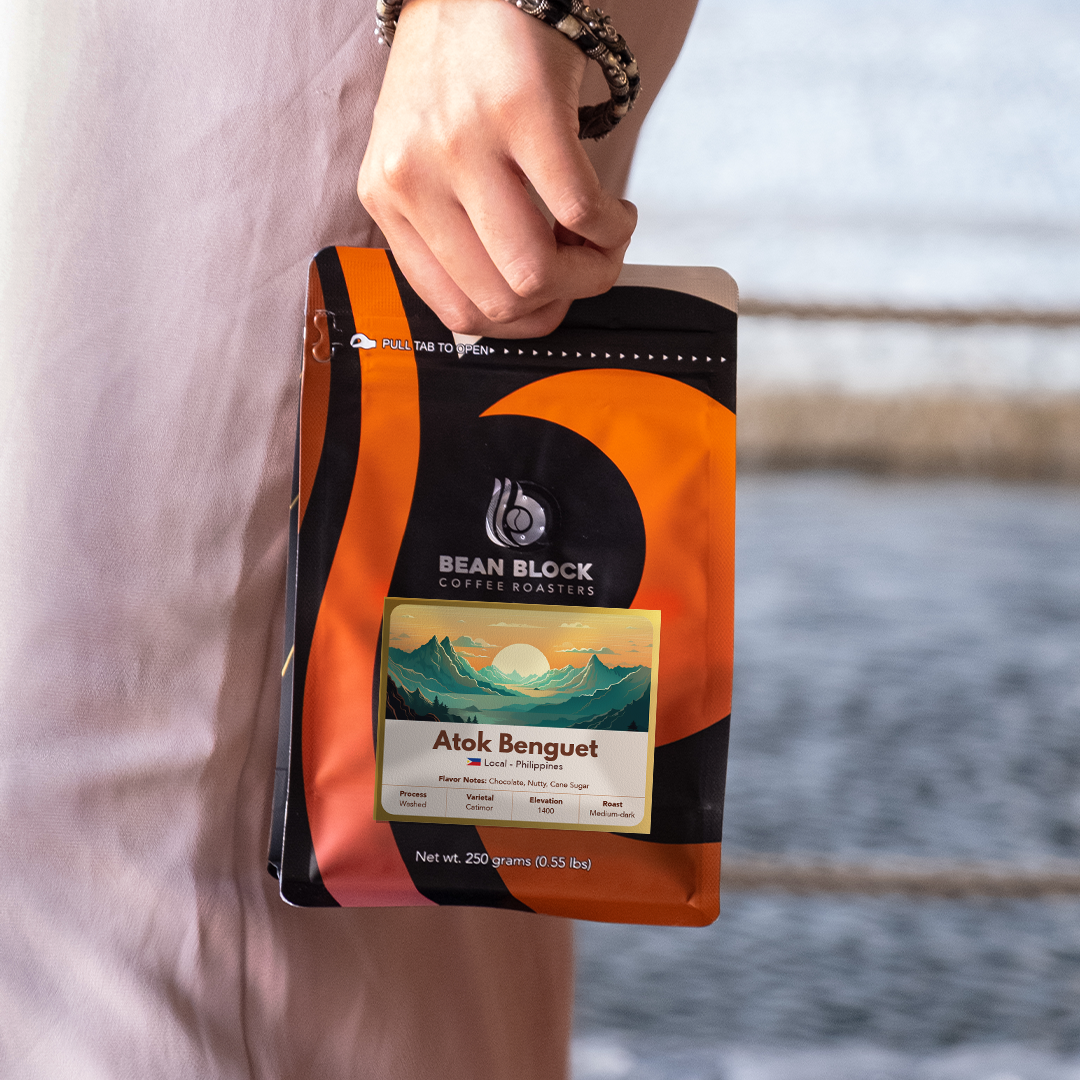 A person in a light pink dress holds a bag of Bean Block Coffee Roasters Atok Benguet coffee against a blurred outdoor background of water and a railing.