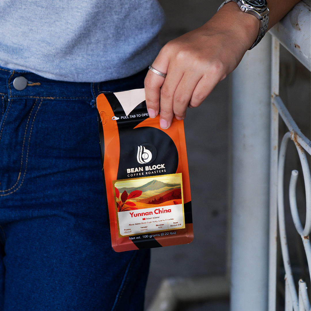 A person in a gray t-shirt and blue jeans holds a pouch of Bean Block Coffee Roasters Yunnan China coffee against a concrete wall background.