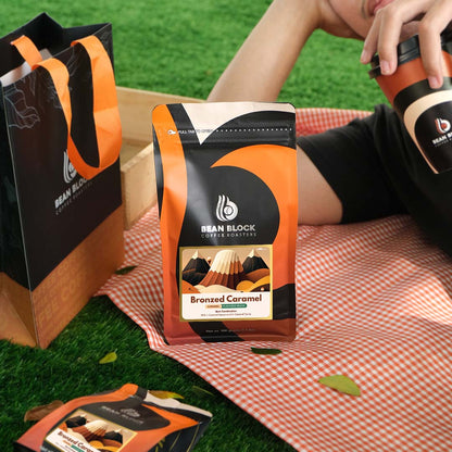 Bean Block coffee roasters 500g Bronzed Caramel flavored coffee package on a picnic blanket with a person holding a coffee cup.