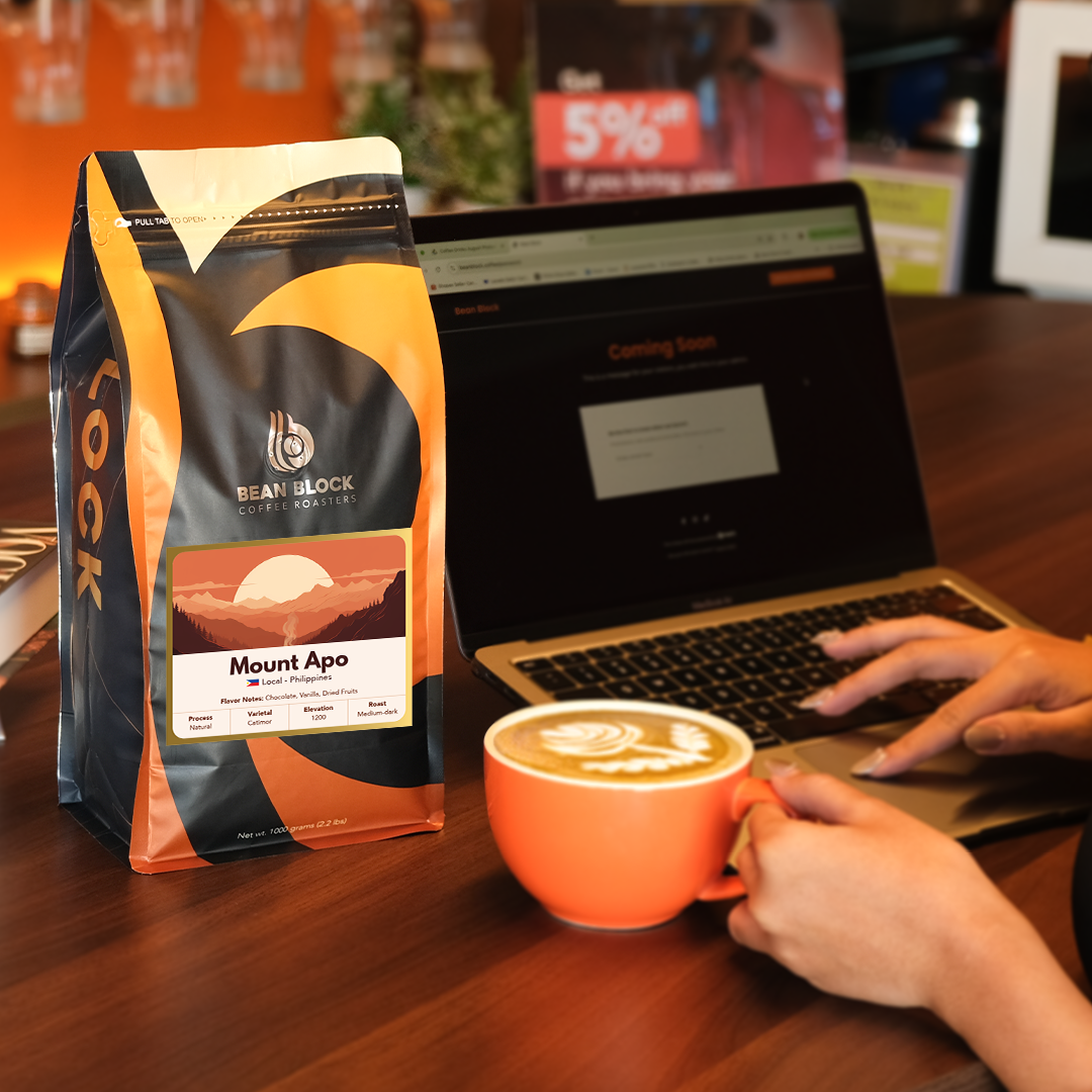 Person holding a coffee cup with a Bean Block Coffee Roasters Mount Apo coffee bag on a table while using a laptop. 1kg of Bean Block Coffee Roasters Coffee Bean pouch bag