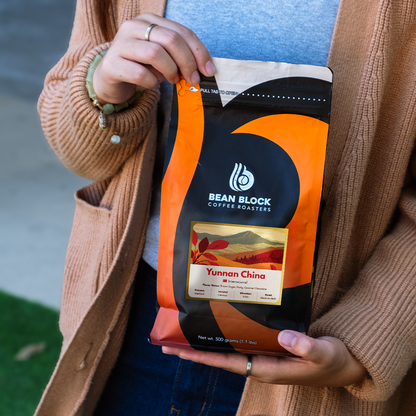 A person in a beige cardigan holds a bag of Bean Block Coffee Roasters Yunnan China coffee. The bag is black and orange with a clear window showing the dark roasted coffee beans inside.