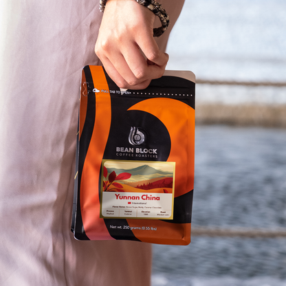 A person holding a stand-up pouch of Bean Block Coffee Roasters Yunnan China coffee against a blurred outdoor background.